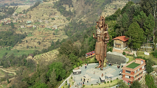 Aerial shot circles around the massive Kailashnath Mahadev Statue, the world's tallest Hindu Shiva statue. Terraced farms and green hills surround the statue in the background.